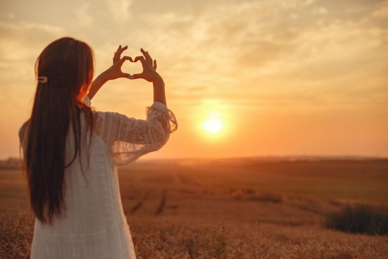 A woman stands in a field making a heart shape with her hands against a beautiful sunset.